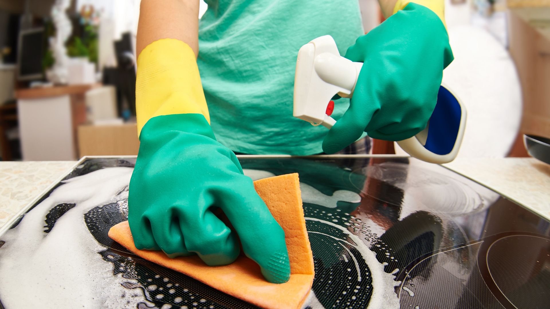 Hands in green gloves cleaning a stovetop with spray and orange sponge